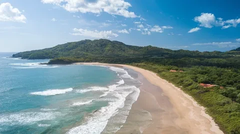 Luftaufnahme eines Sandstrandes an der Pazifikküste v Costa Rica, umgeben von türkisfarbenem Wasser und üppig grünen Hügeln.